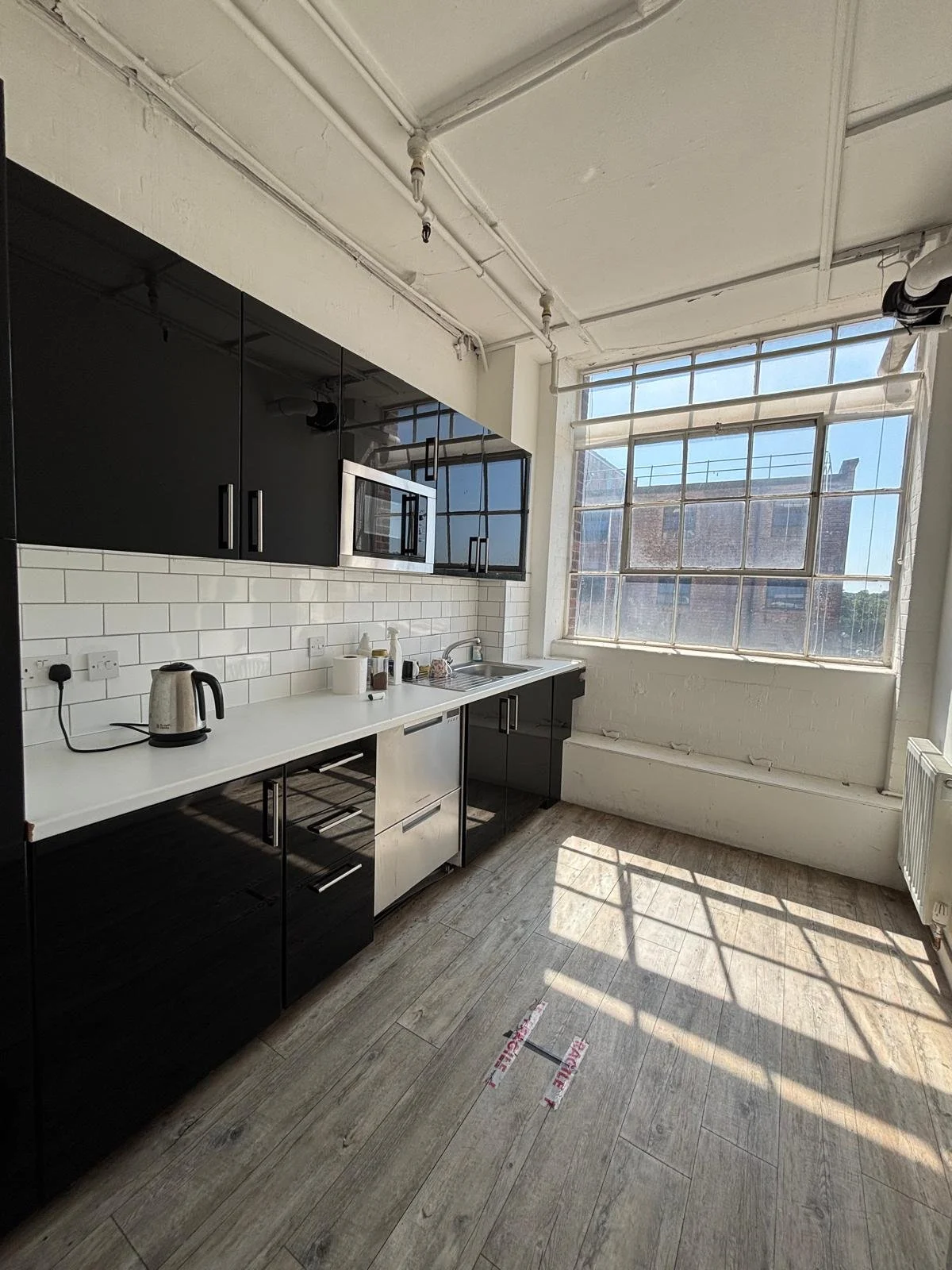 Modern kitchen with black cabinets, white countertop, white subway tile backsplash, stainless steel appliances, kettle, and large window with a city view, wooden flooring, and exposed ceiling pipes.