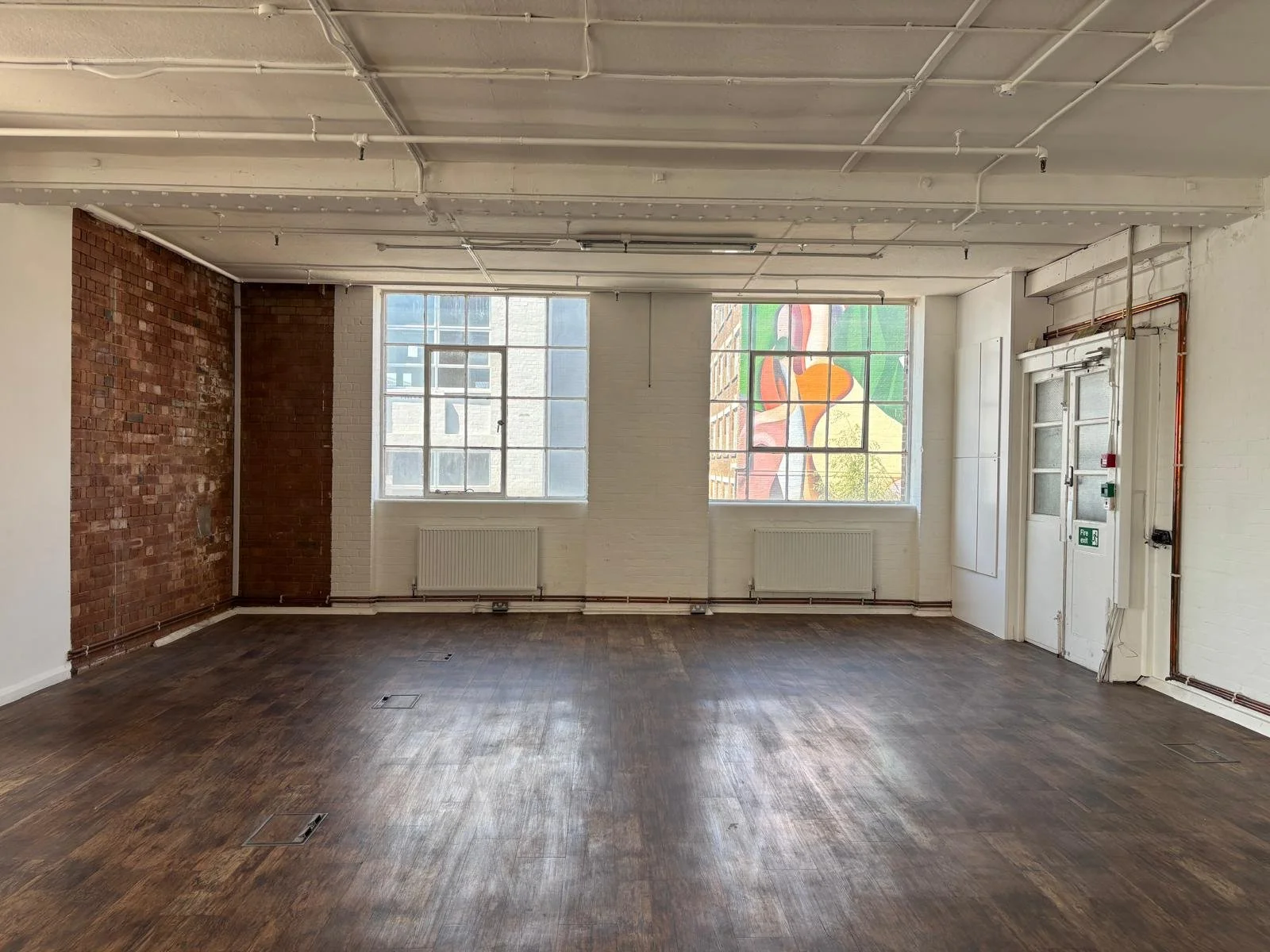 An empty room with white brick walls, a wooden floor, large windows, exposed ceiling pipes, and a fire exit door.
