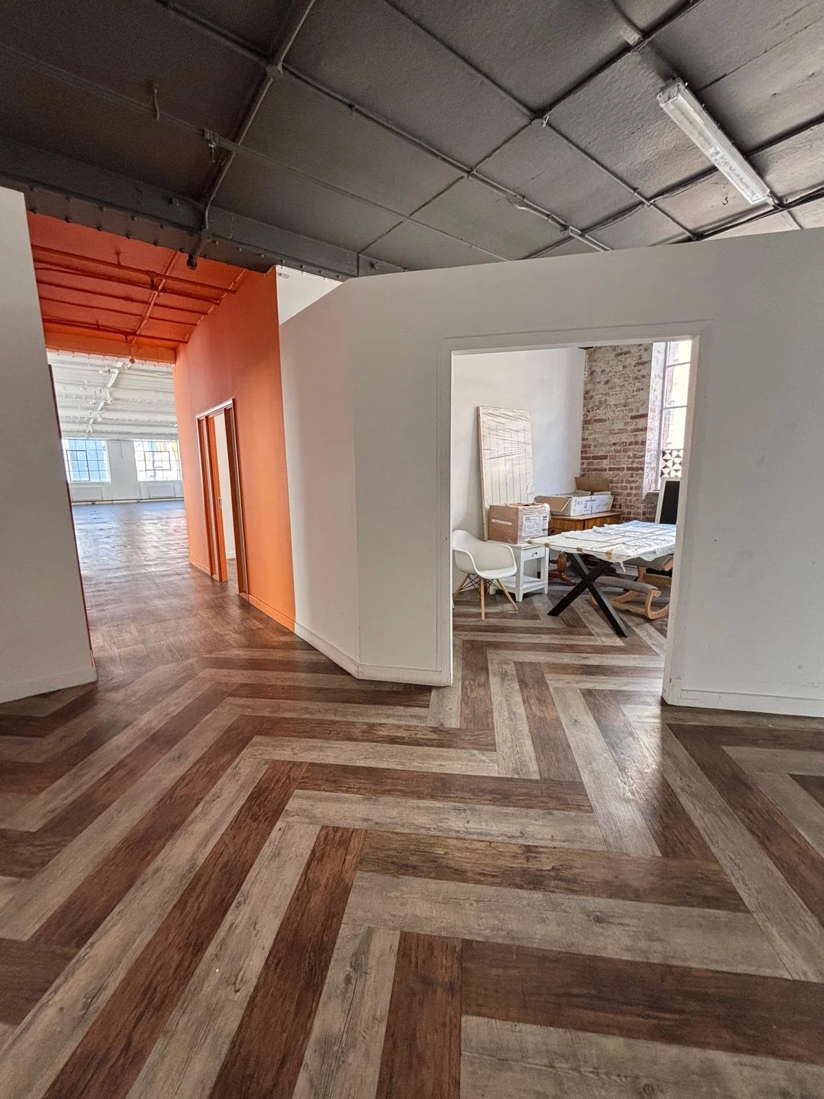Interior of a modern industrial-style office or studio space with chevron wood flooring, white and orange painted walls, exposed ceiling pipes, and large windows letting in natural light. There is a small room with equipment and supplies, and a corridor leading to a larger open area.