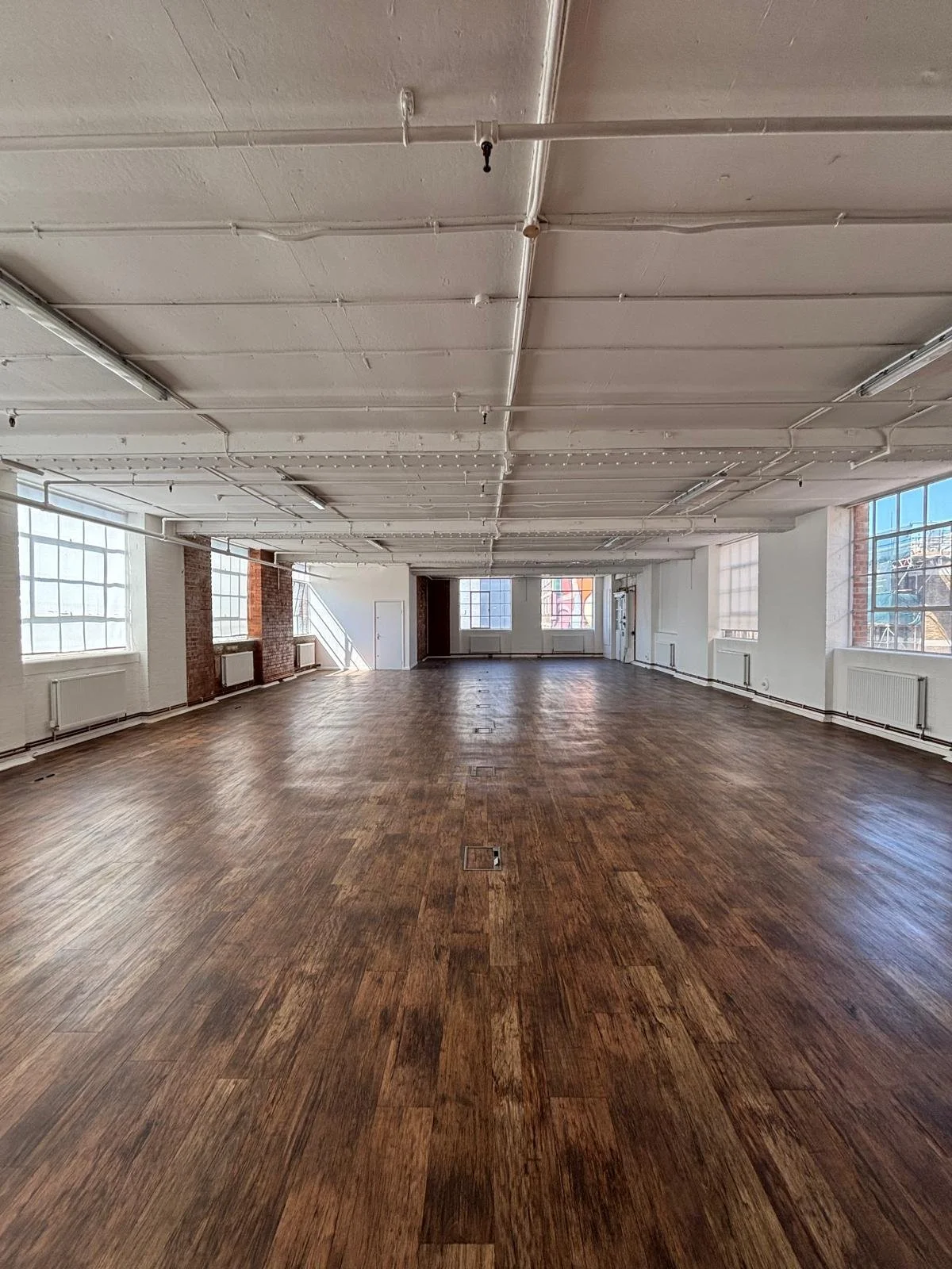 Large, empty industrial-style room with wood flooring, white walls, large windows, and exposed ceiling pipes.