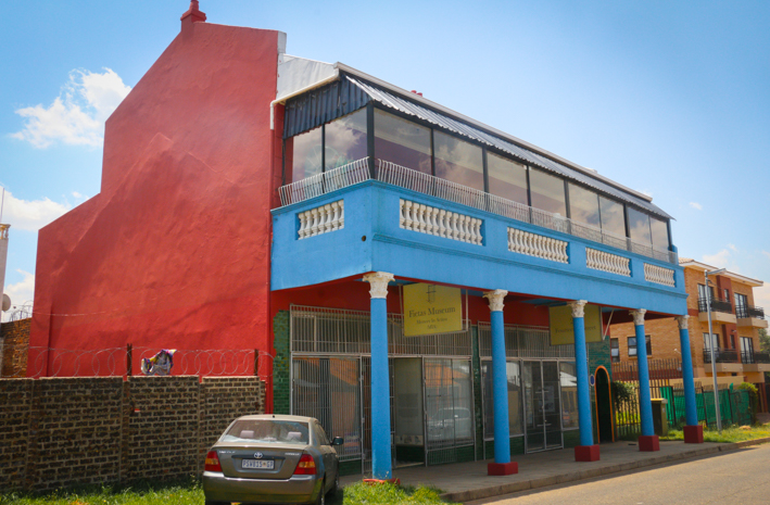 A two-story building with bright red and blue exterior colors. The upper level has large glass windows and a balcony with a metal railing, sheltered by an awning. The ground floor has a gated entrance and blue supporting columns, with a car parked in front on the street.