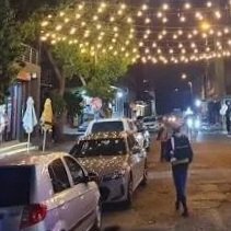 Street scene with parked cars, string lights overhead, and a person walking at night.