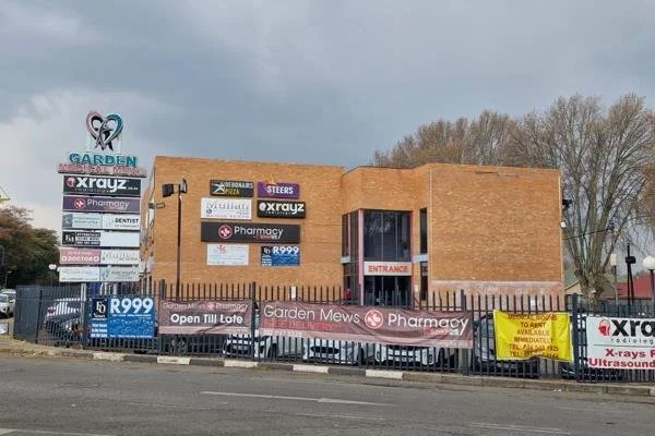 A two-story brick building with signs for various businesses including a pharmacy, X-ray, and dentistry, surrounded by a black metal fence and a sidewalk, under a cloudy sky, with trees in the background.