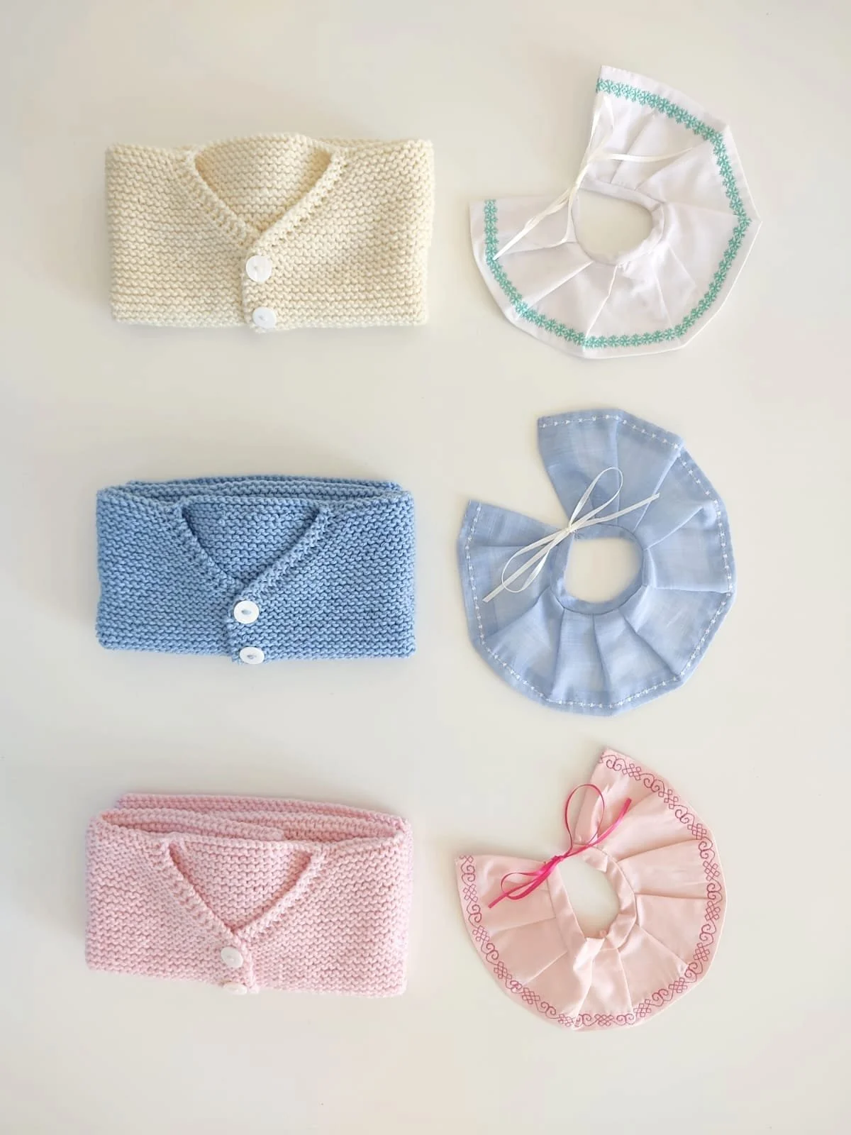 Tre golfini neonato piegati accanto a tre colletti plissé ricamati in bianco, azzurro e rosa con nastrino