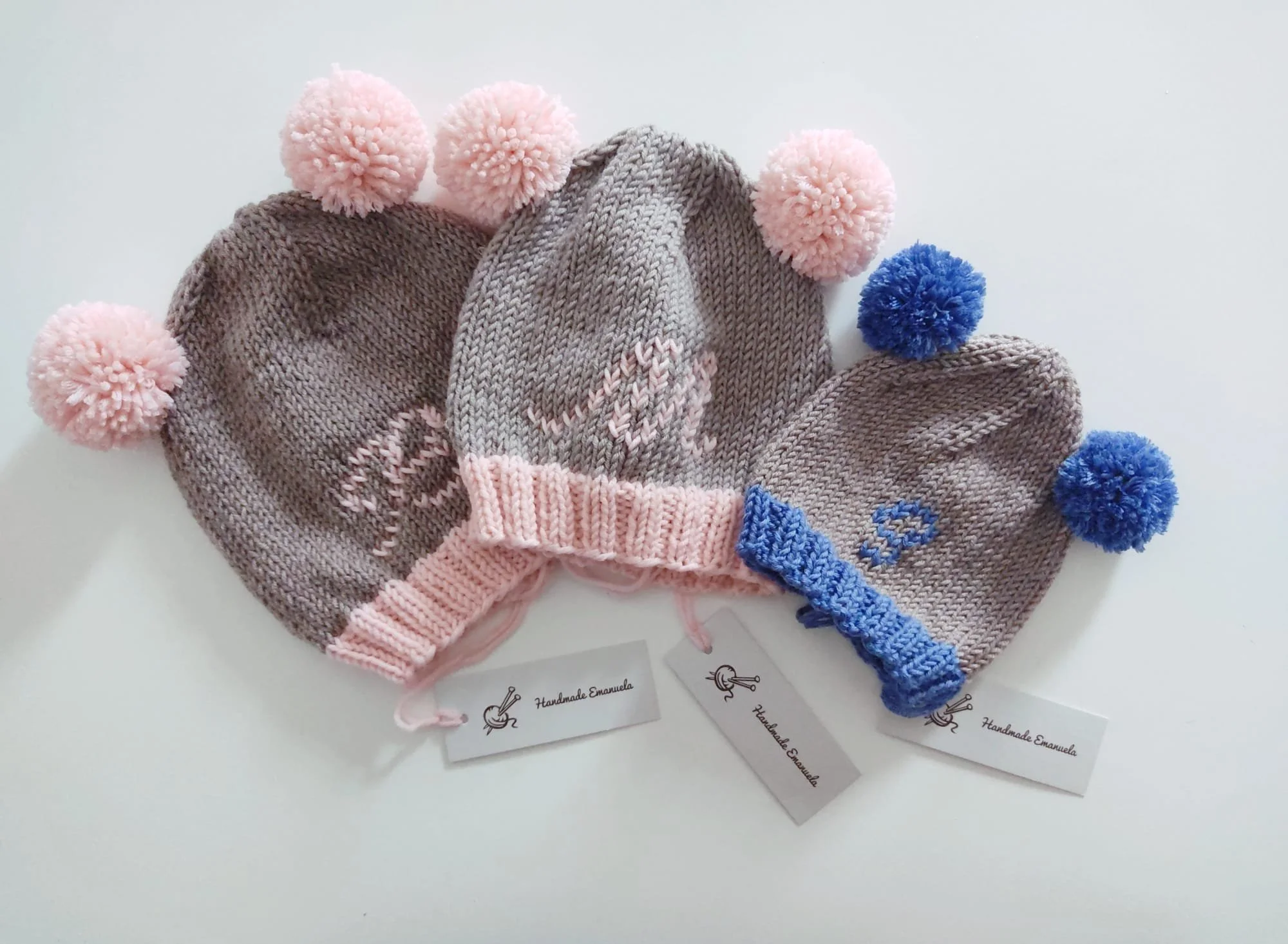 Tre cappelli in maglia grigio tortora con iniziale ricamata, pompon rosa o azzurri e bordo a coste, con cartellino