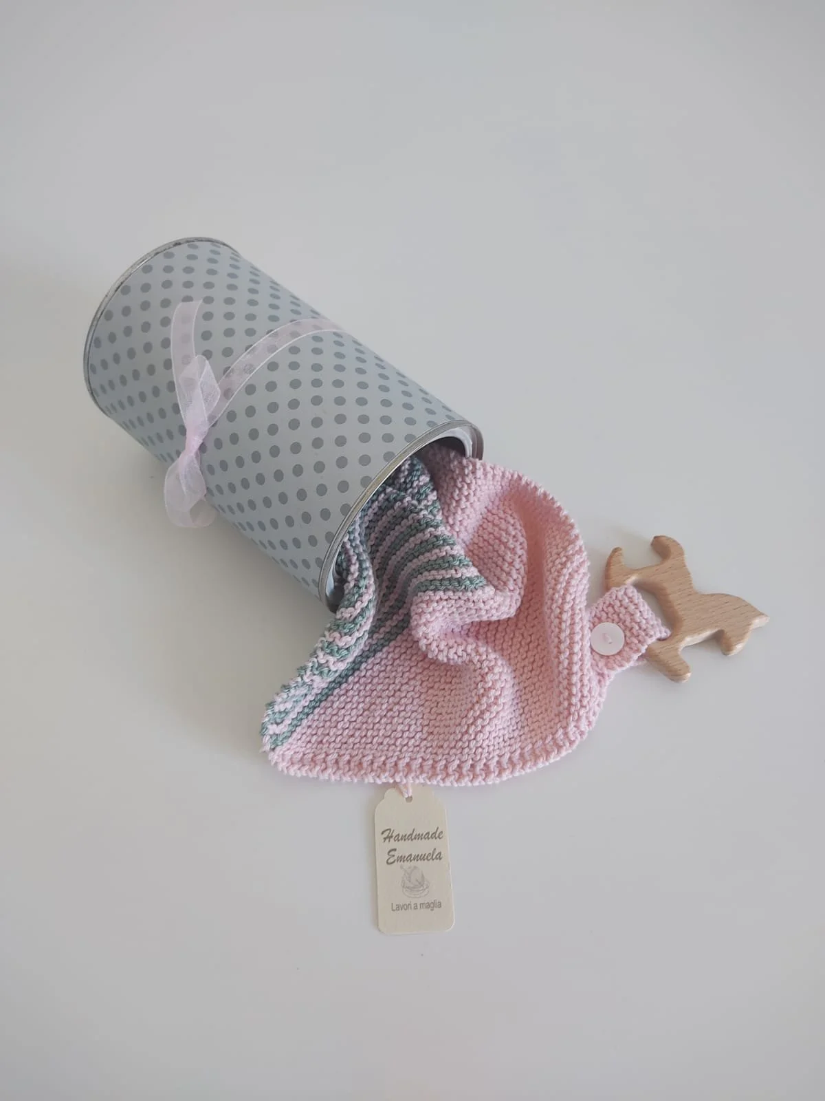 Doudou in maglia rosa con righe verdi che esce da un barattolo grigio a pois, con giochino in legno e cartellino