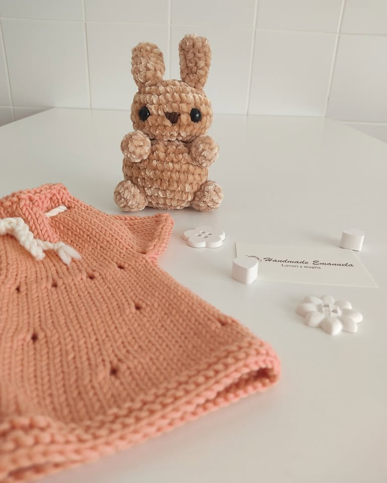 Coniglietto di ciniglia beige accanto a un abitino in maglia color pesca, con cartellino e bottoni bianchi