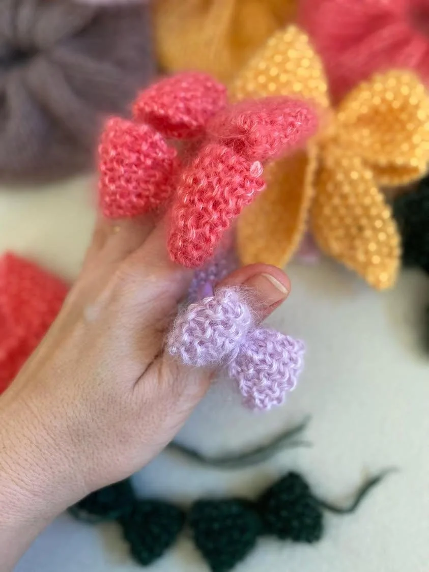 Mano che regge fiocchetti in mohair lavorati a maglia, rosa corallo, arancio e lilla, tra altri colori sullo sfondo