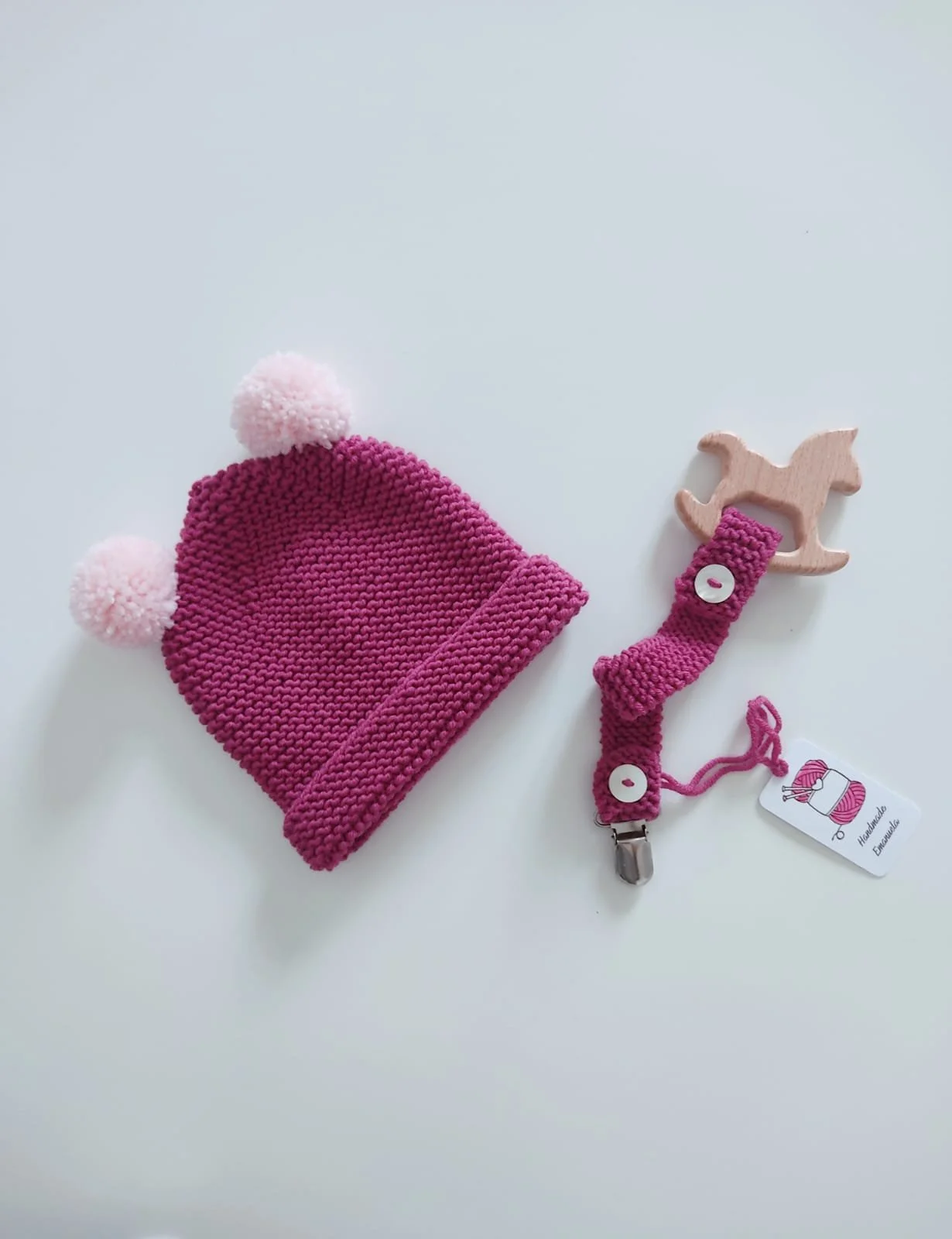 Portaciuccio in maglia fucsia con giochino in legno a cavallino a dondolo, accanto a un cappellino fucsia con due pompon rosa