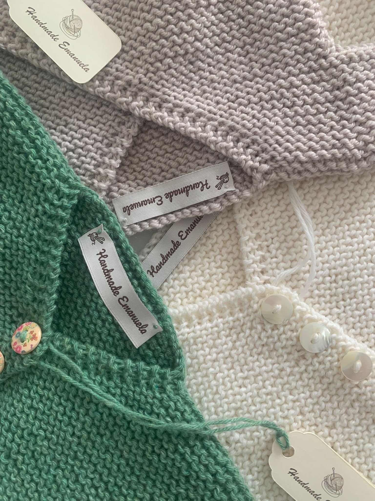 Dettaglio di tre golfini a maglia verde, tortora e panna, con etichette Handmade Emanuela e bottoni in madreperla e a fiori.