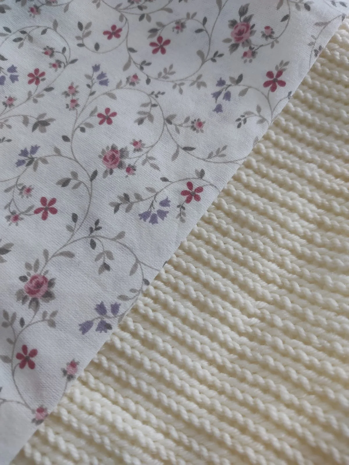 Dettaglio del cotone a fiorellini rosa e lilla accanto a una coperta in maglia color panna