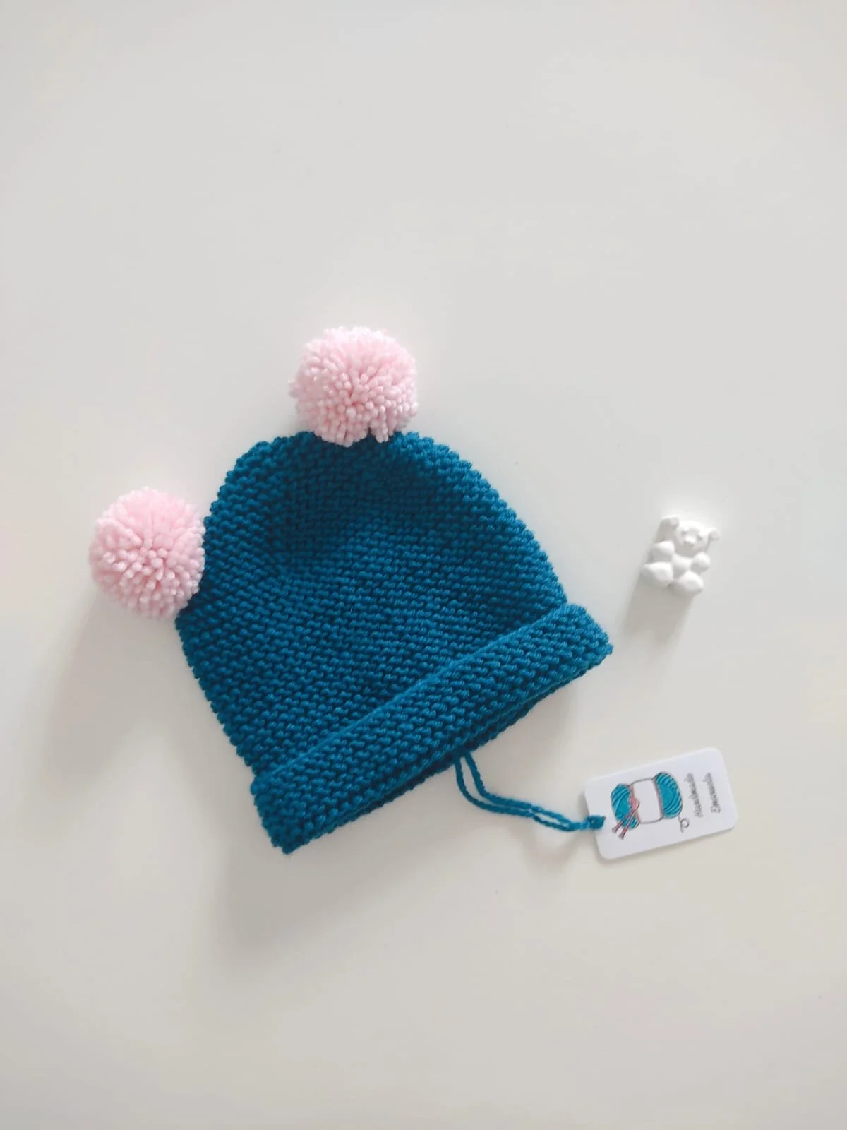 Cappello in maglia blu petrolio con risvolto e due pompon rosa chiaro, con cartellino