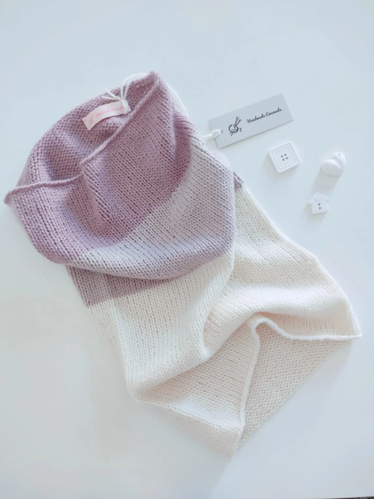 Scaldacollo a maglia bicolore lilla e panna, con etichetta Handmade Emanuela e cartellino grigio con gomitolo.