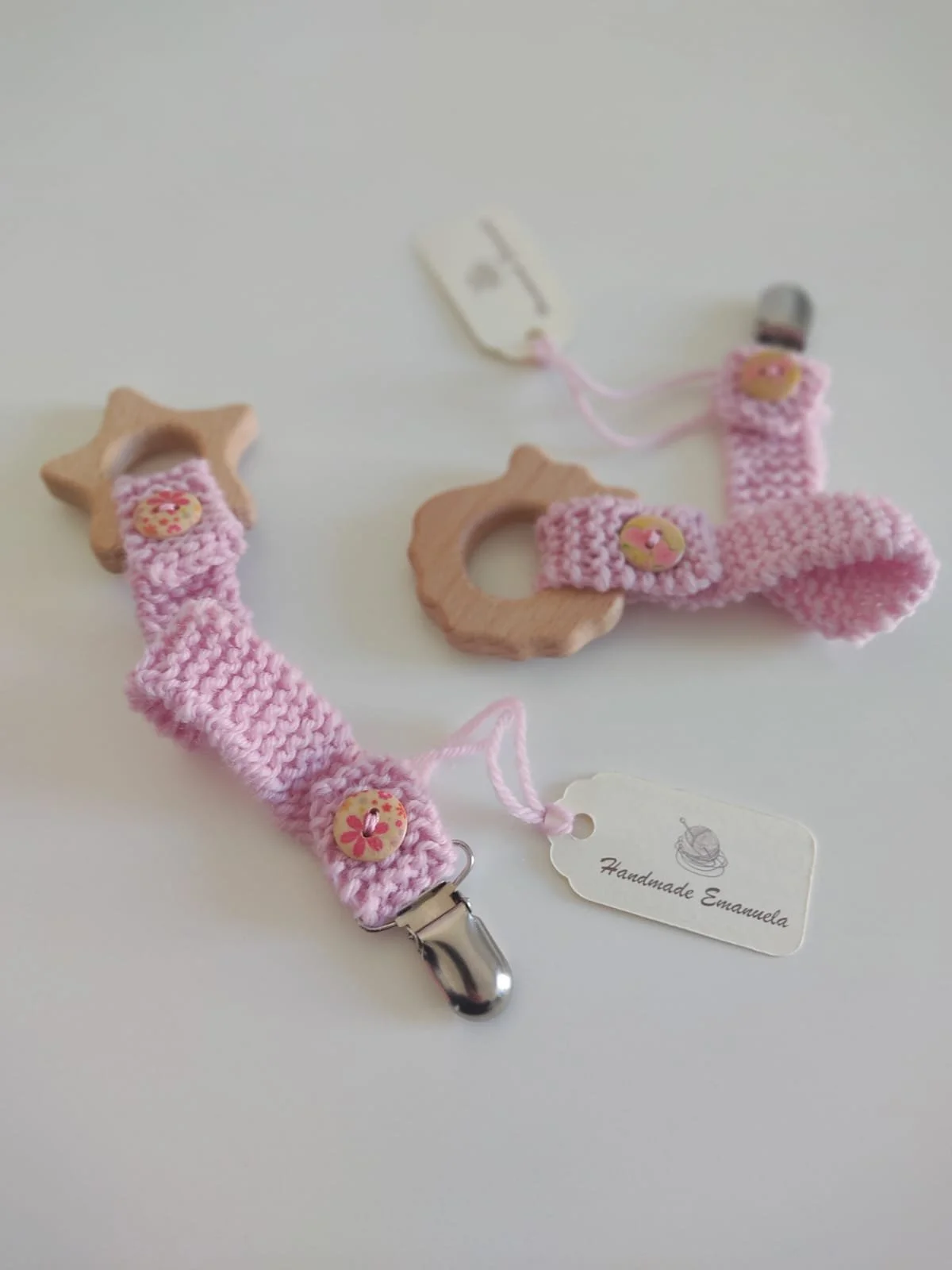 Due portaciuccio in maglia rosa con giochino in legno a stella e a riccio, bottoni in legno a fiorellini e cartellini