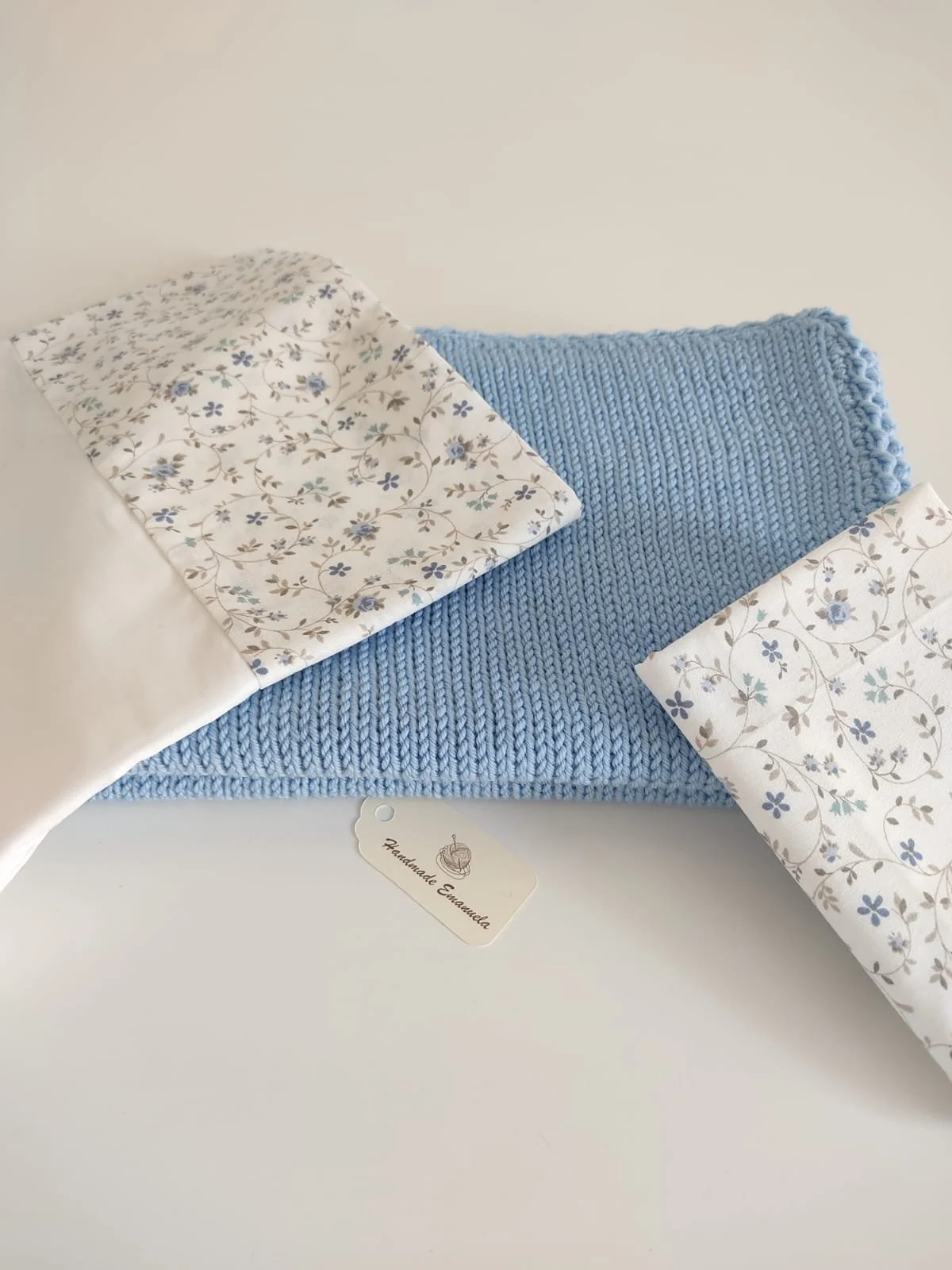 Lenzuolino e federa in cotone bianco a fiorellini azzurri piegati su una coperta in maglia azzurra, con cartellino