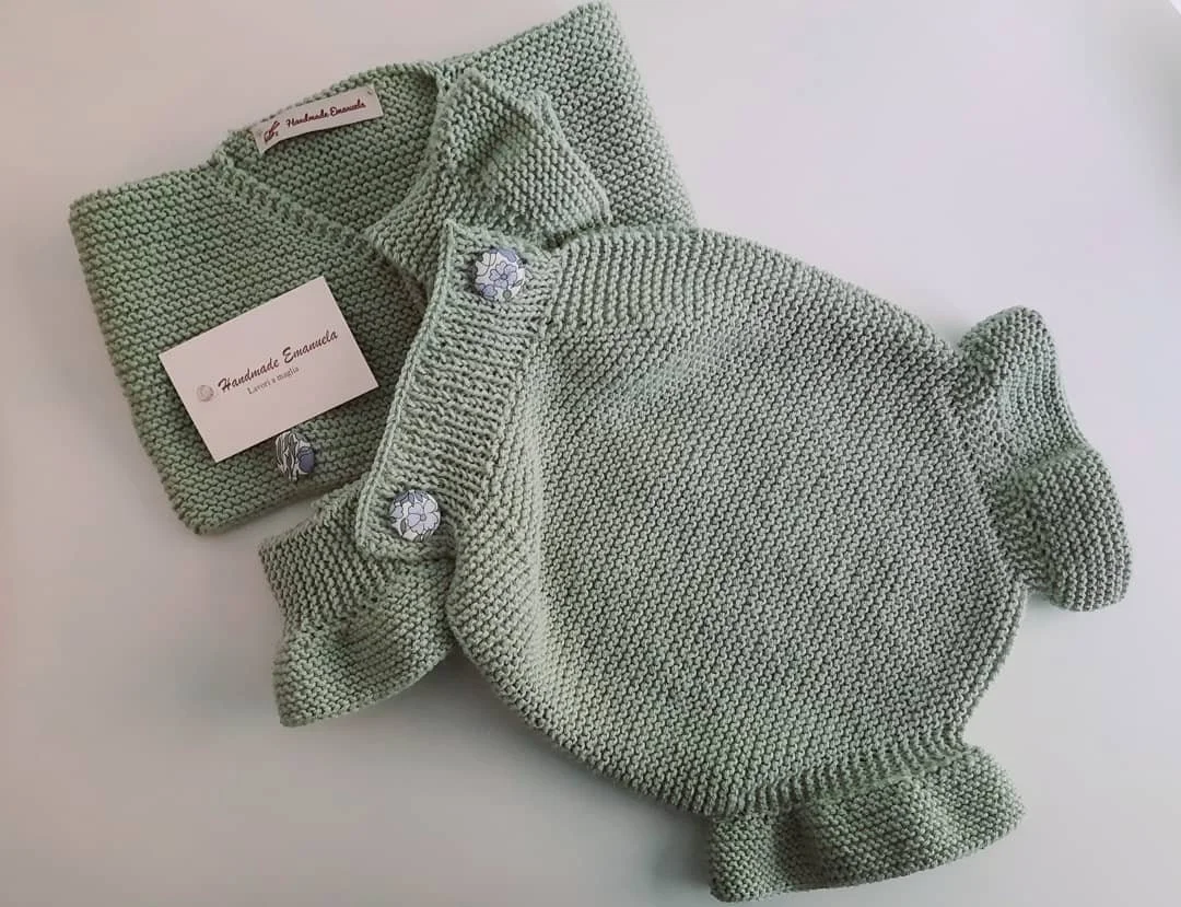 Pagliaccetto a maglia verde salvia con volant sulle gambine e bottoni rivestiti in stoffa a fiori, accanto a un golfino dello stesso colore.