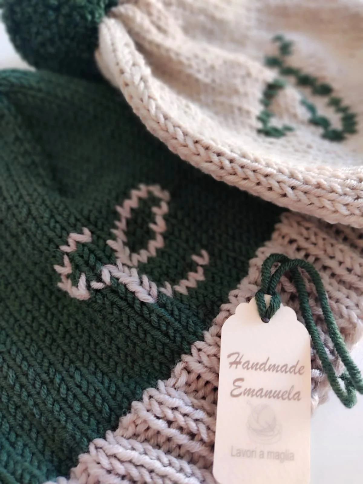 Primo piano di due cappelli in maglia verde scuro e beige con iniziale ricamata a contrasto e cartellino