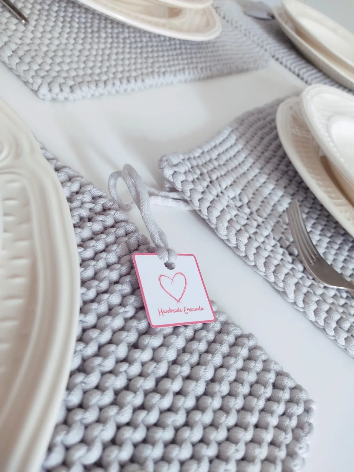 Tovagliette a maglia grigio chiaro con cartellino a cuore Handmade Emanuela, apparecchiate con piatti bianchi e forchetta.