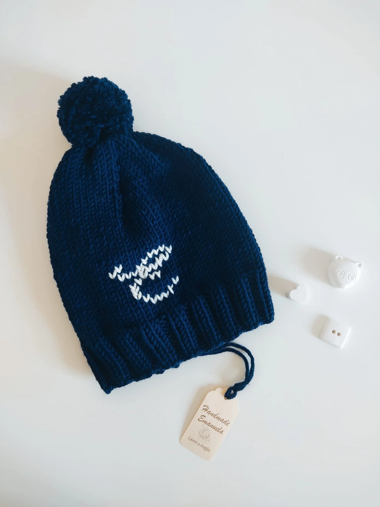 Cappello in maglia blu scuro con pompon dello stesso colore, iniziale ricamata bianca e bordo a coste