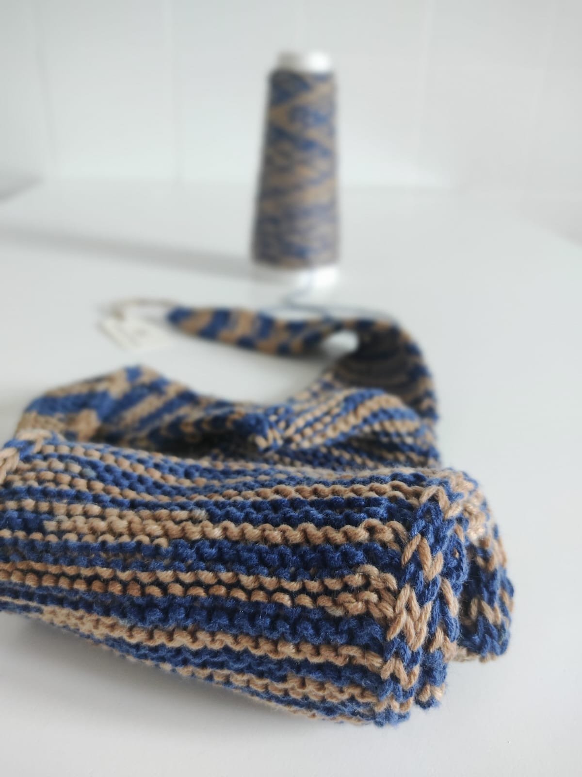 Dettaglio di una sciarpina in maglia a righe blu e cammello, con rocca di filato sullo sfondo