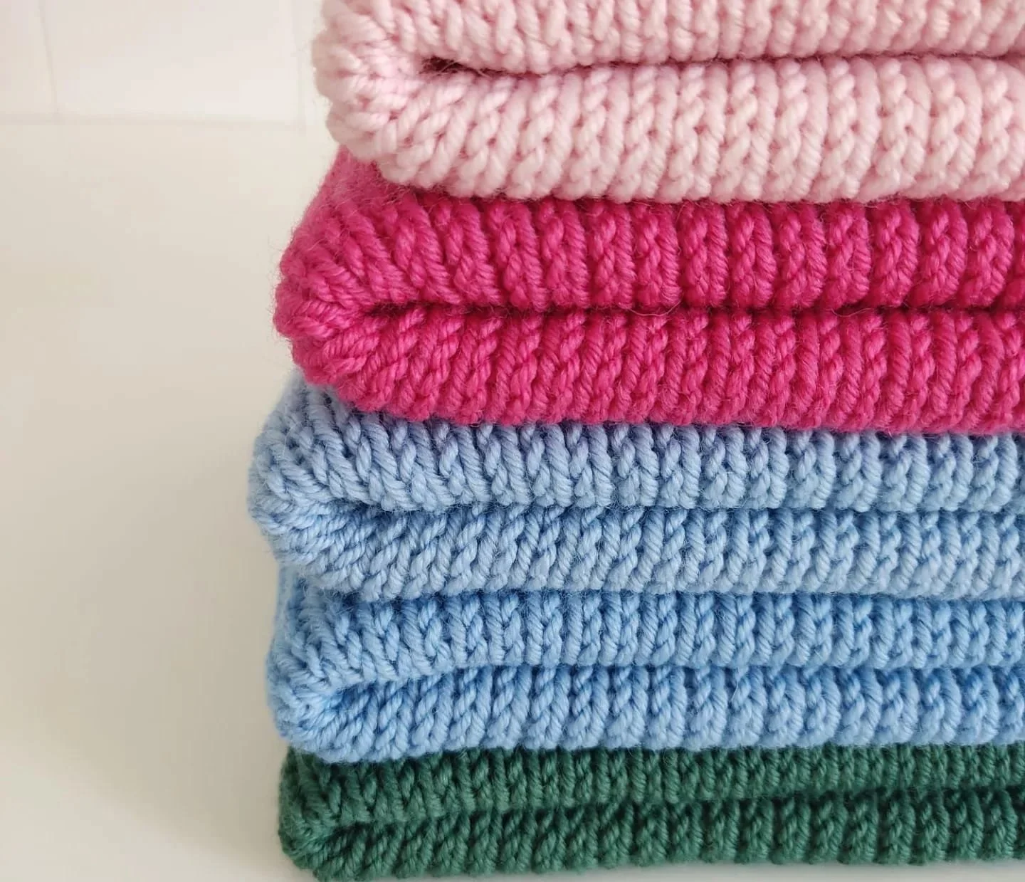 Pila di lavori a maglia piegati in rosa chiaro, fucsia, azzurro e verde scuro.