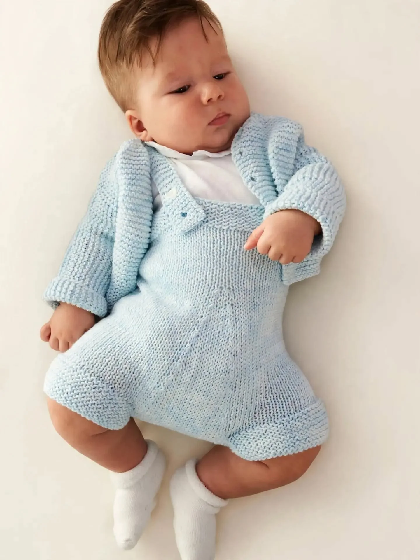 Bambino piccolo sdraiato che indossa una salopette a maglia azzurra con golfino coordinato, camicina bianca e calzini bianchi.