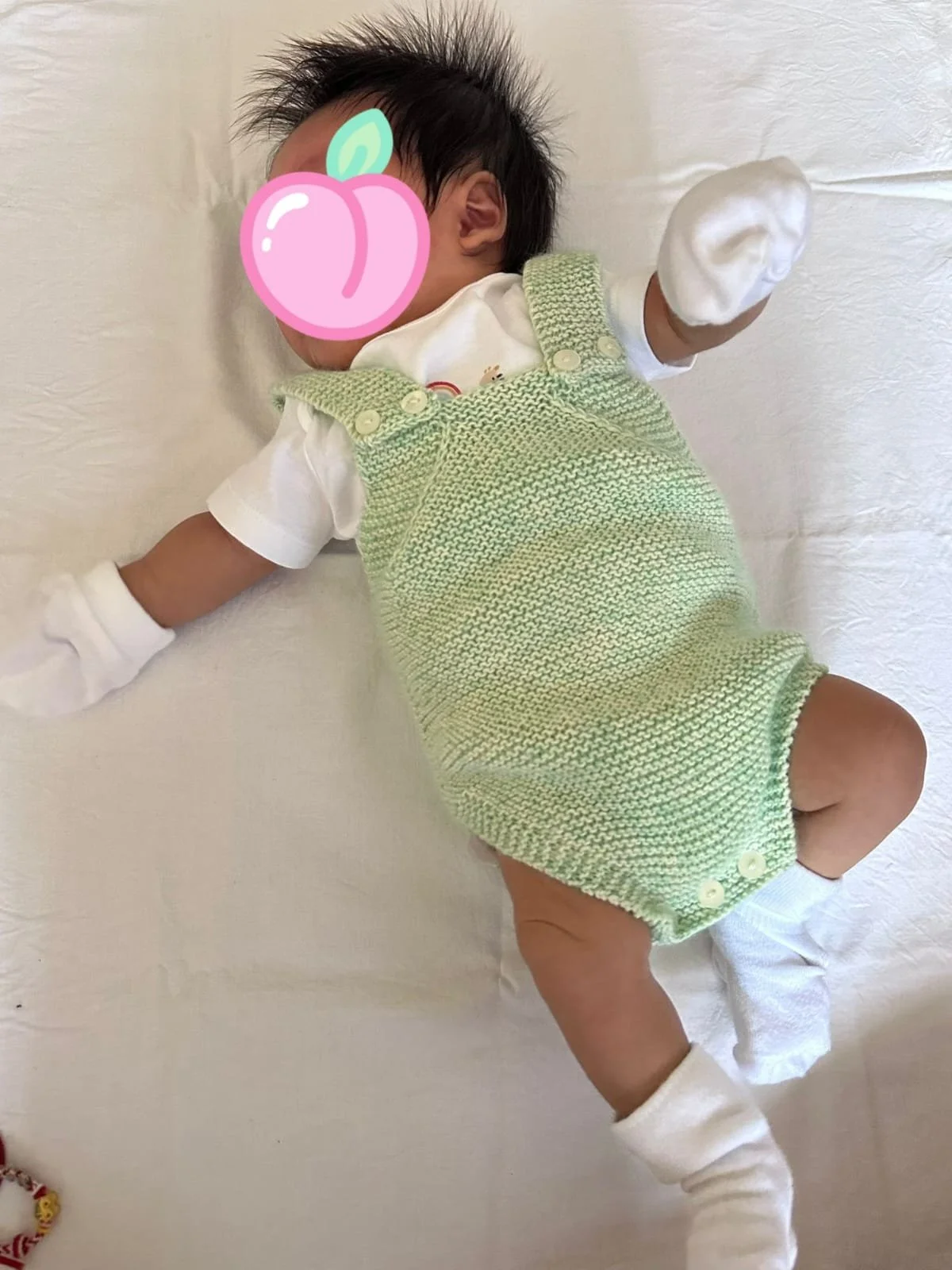 Neonato disteso su un lenzuolo bianco con un pagliaccetto in maglia verde menta con bretelle e bottoncini