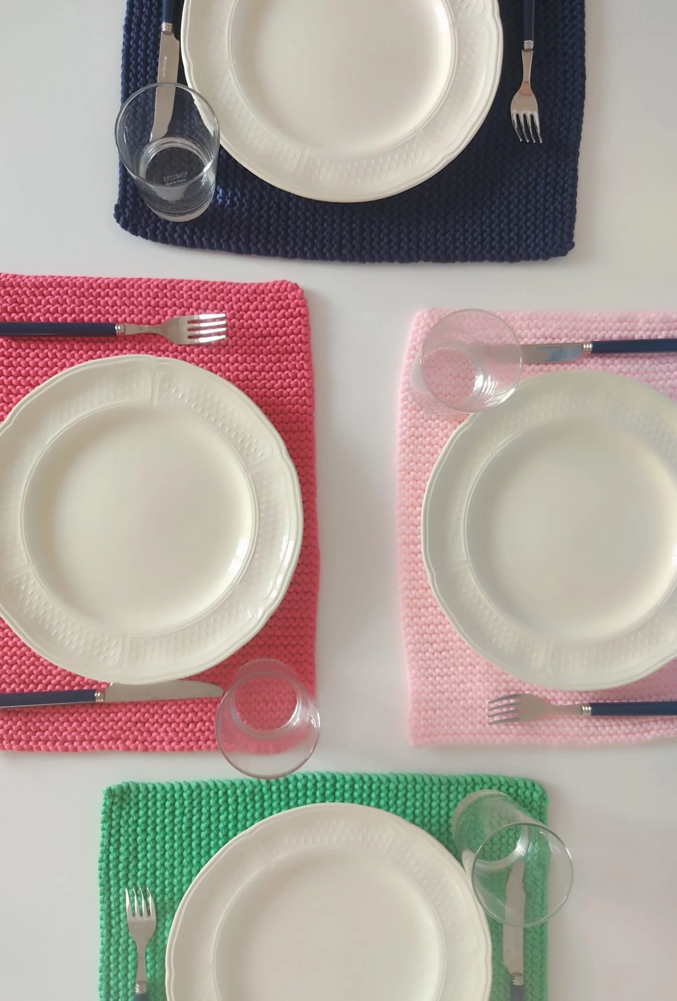 Quattro tovagliette americane a maglia in blu, corallo, rosa e verde, con piatti bianchi, bicchieri e posate.