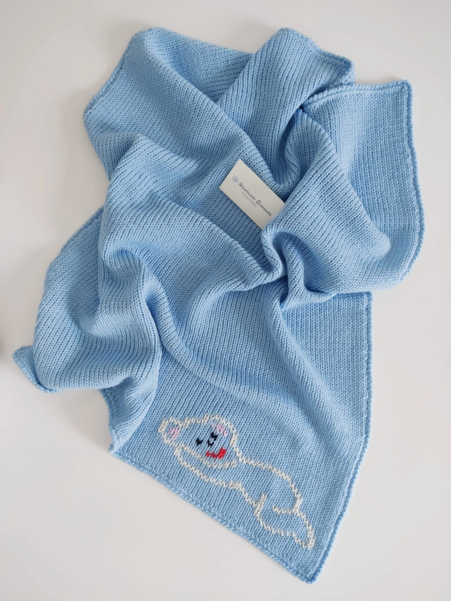 Copertina a maglia azzurra con un piccolo animaletto stilizzato con occhi chiusi lavorato in bianco e rosa.