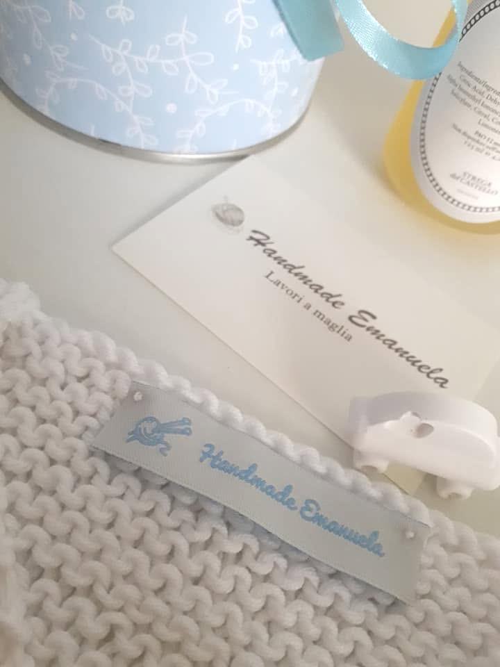 Etichetta Handmade Emanuela cucita su un lavoro a maglia bianco, con cartoncino del brand e una scatola azzurra sullo sfondo.