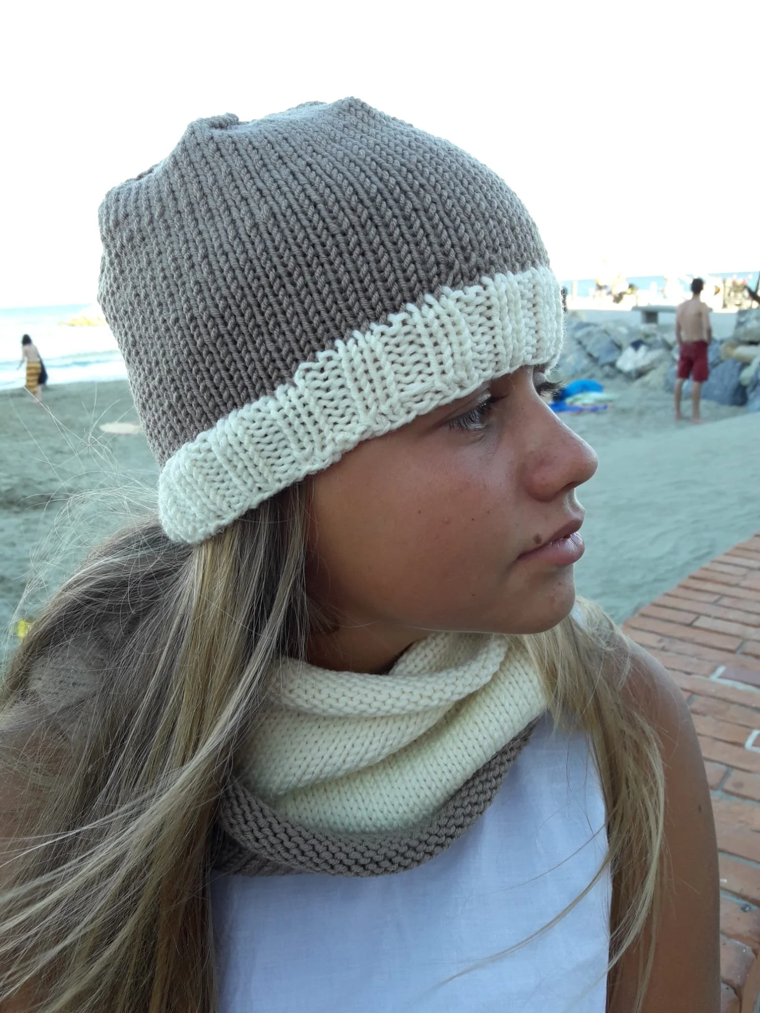 Ragazza di profilo con cappello in maglia beige con bordo crema e scaldacollo crema e beige, sulla spiaggia