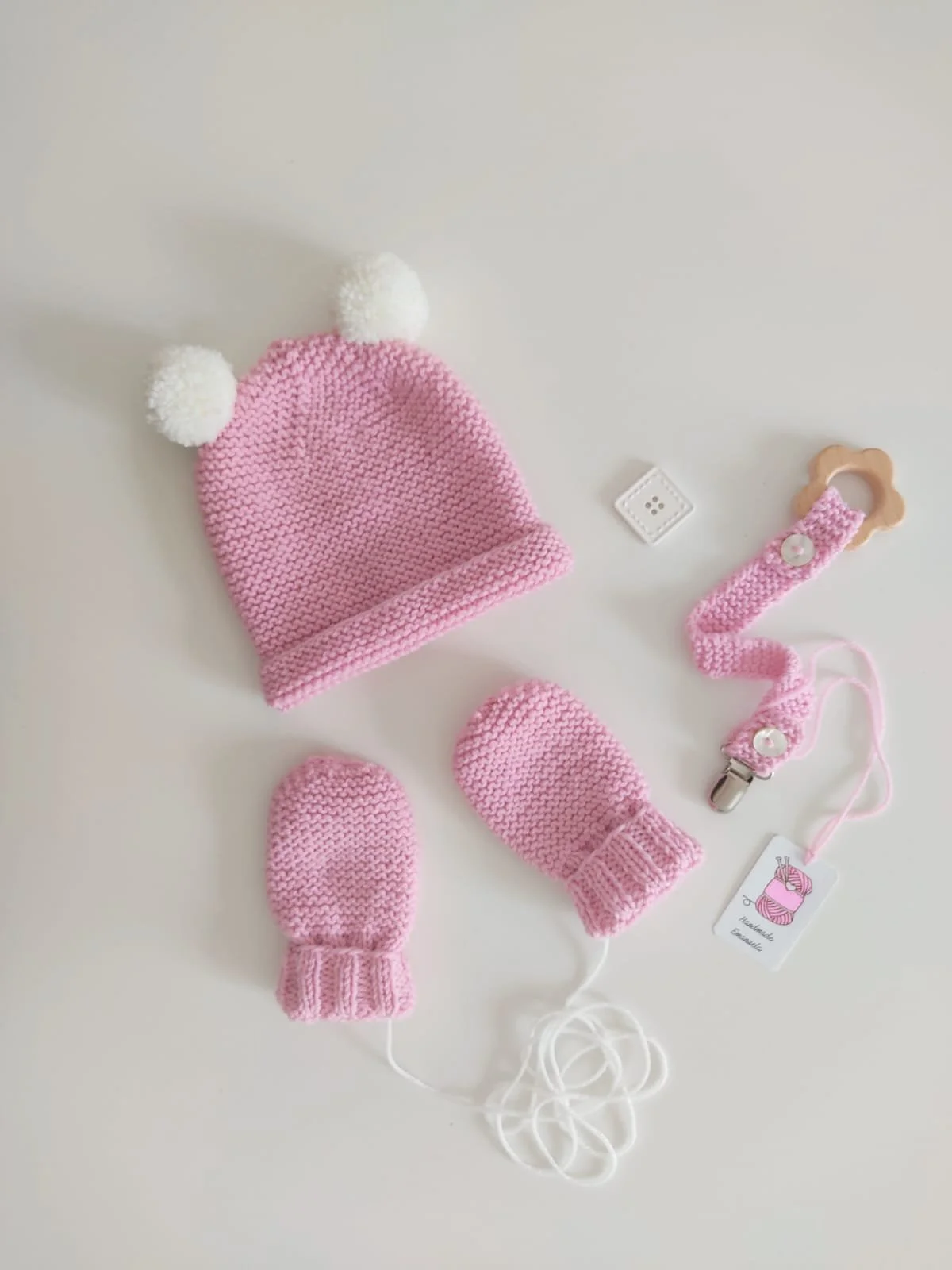 Moffole rosa con laccetto bianco accanto a un cappellino rosa con due pompon crema e a un fermaciuccio abbinato