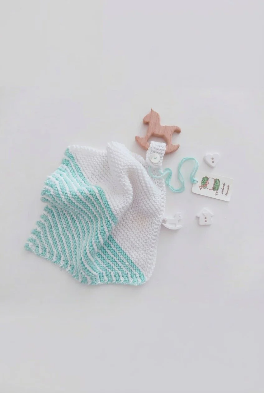 Doudou in maglia bianca con righe verde acqua, giochino in legno a forma di cavallino a dondolo e bottoni bianchi