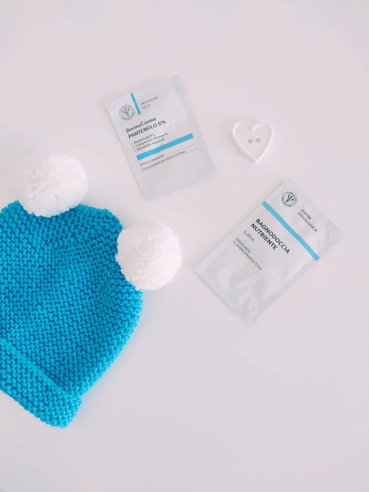 Cappello in maglia turchese con risvolto e due pompon bianchi, accanto a un bottone a forma di cuore