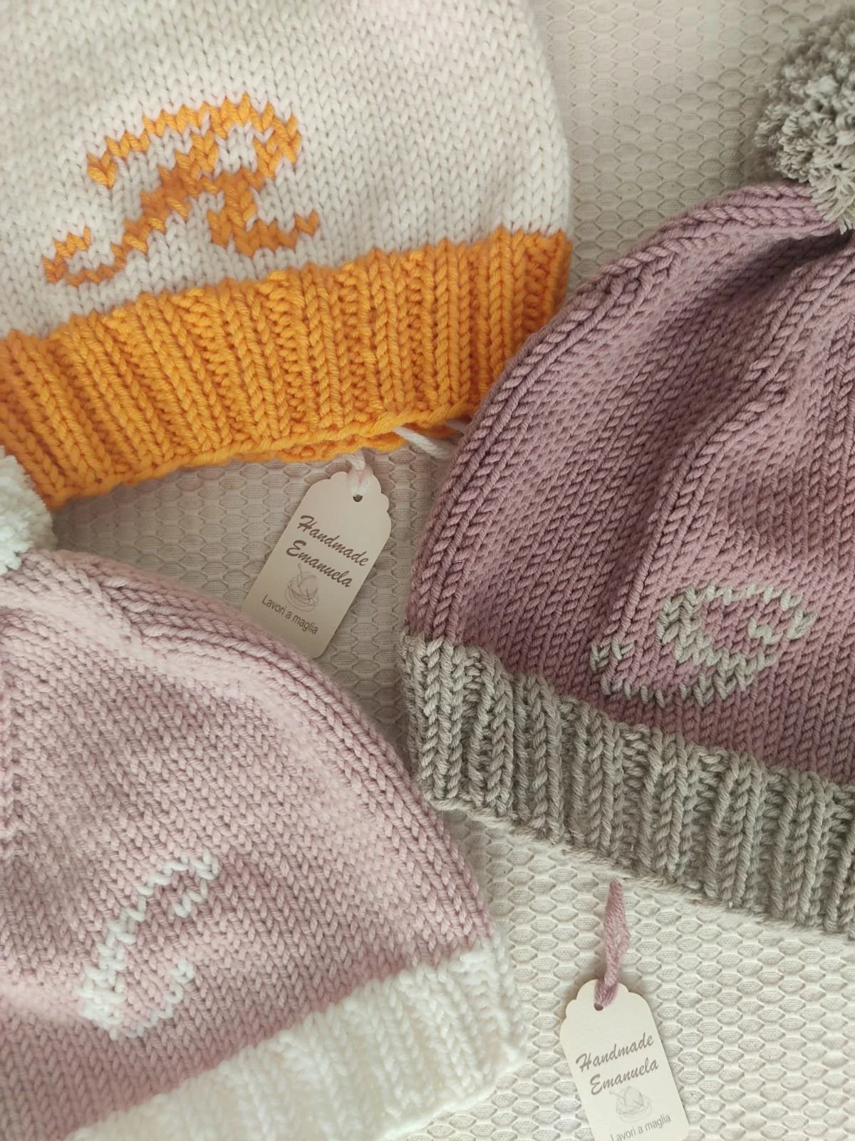 Primo piano di tre cappelli in maglia con iniziale ricamata, nei toni del rosa, tortora e arancione, con cartellini
