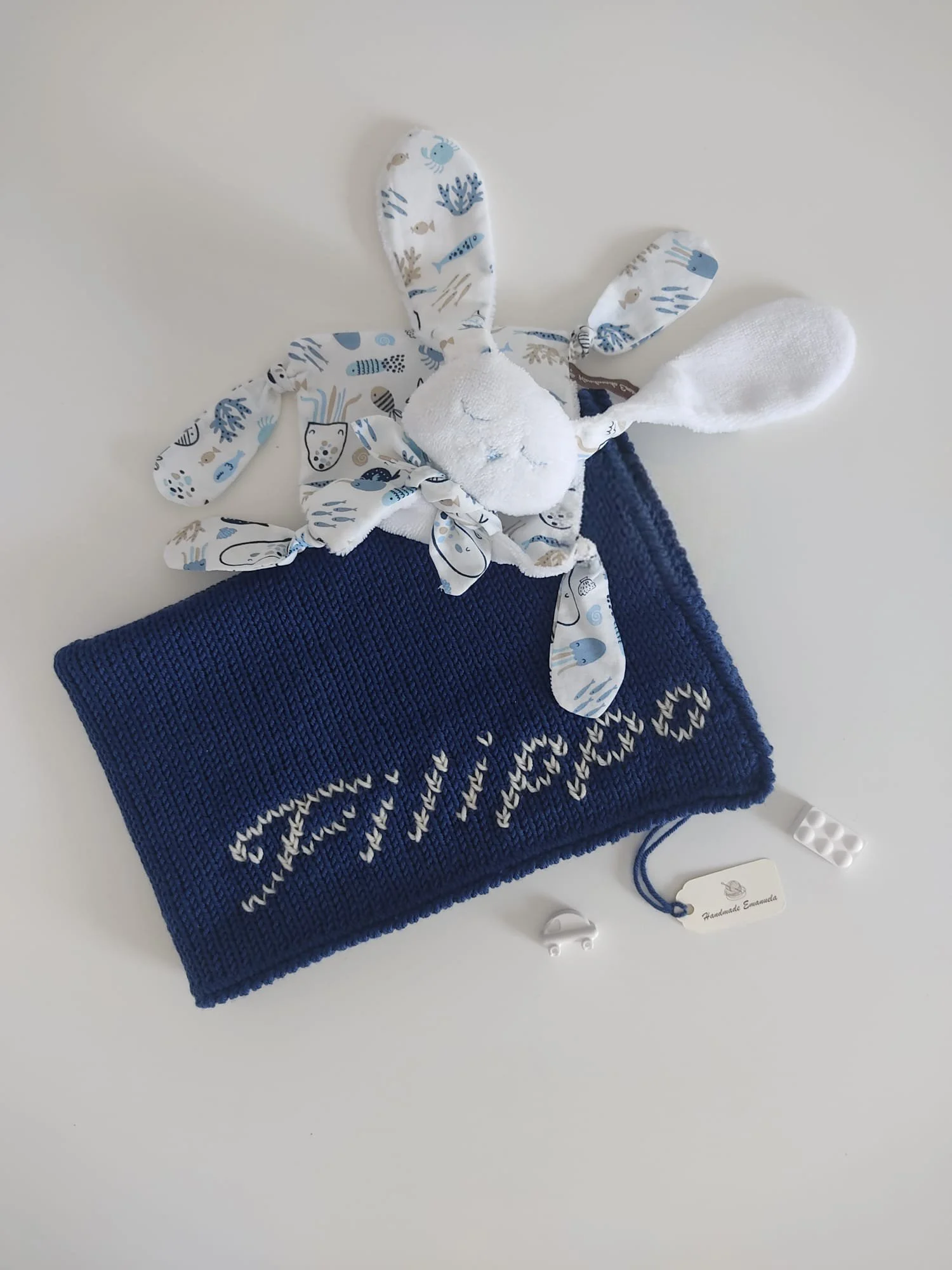 Doudou coniglietto in spugna bianca con orecchie a fantasia di animali marini, su una copertina in maglia blu con una scritta chiara