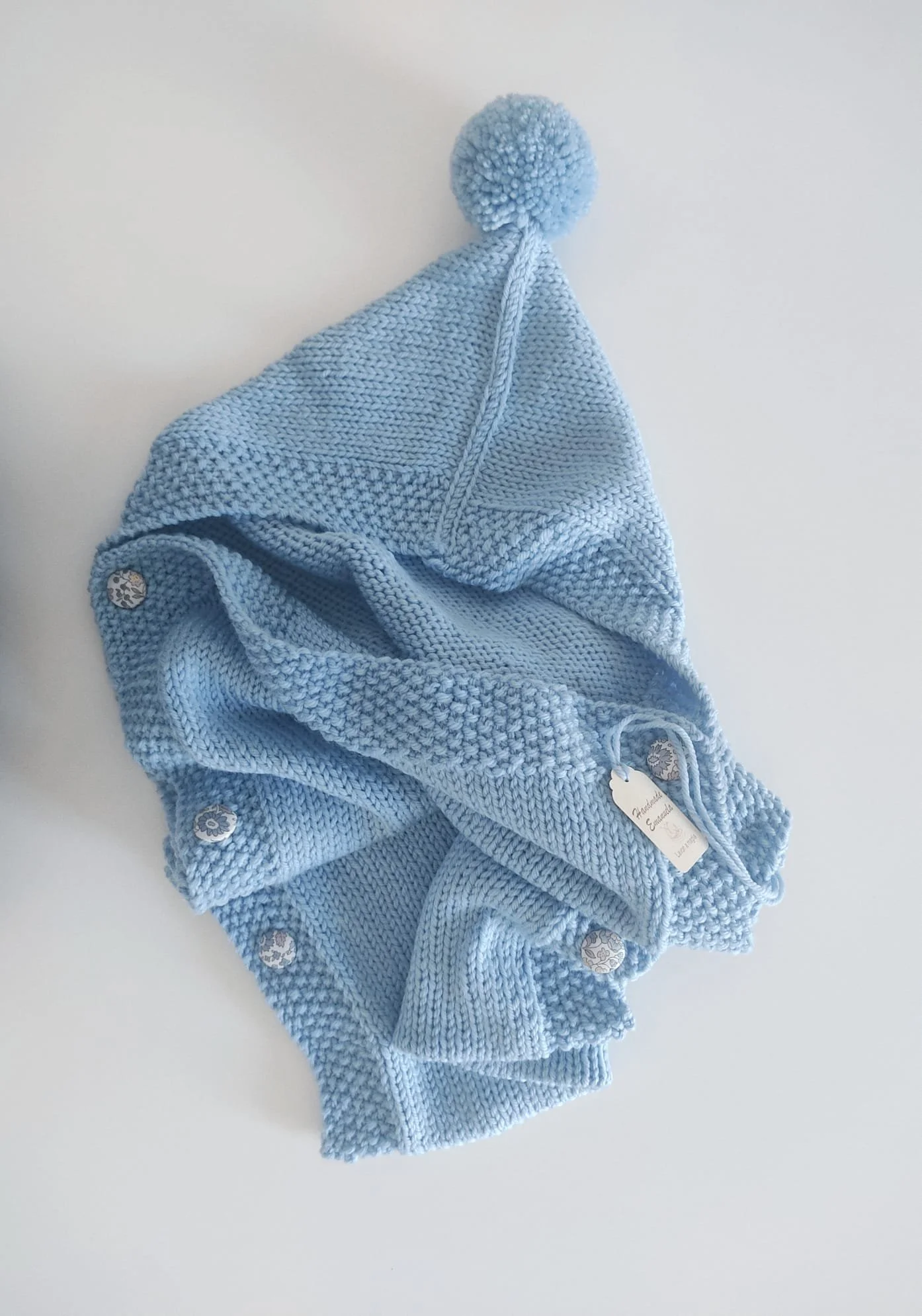 Sacco nanna azzurro in maglia con cappuccio e pon pon, bottoni rivestiti e cartellino, appoggiato su fondo chiaro