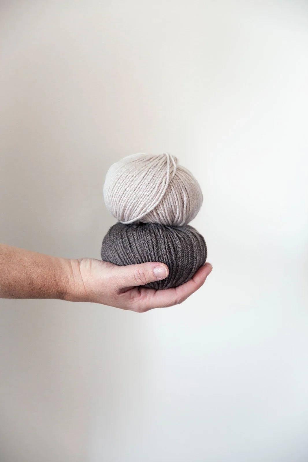Una mano regge due gomitoli di lana, uno beige chiaro sopra e uno grigio scuro sotto, su sfondo chiaro