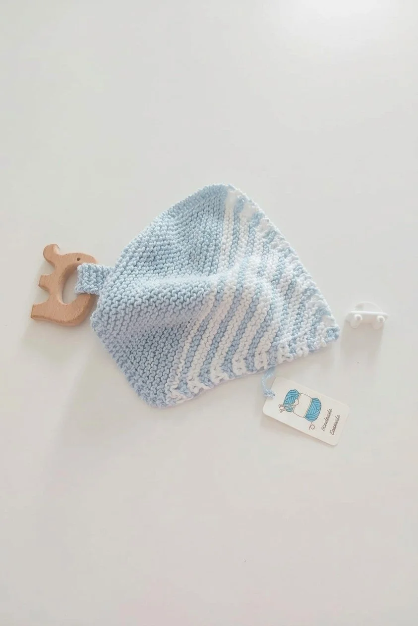 Doudou in maglia azzurra con righe bianche e giochino in legno, accanto a un cartellino di cartoncino