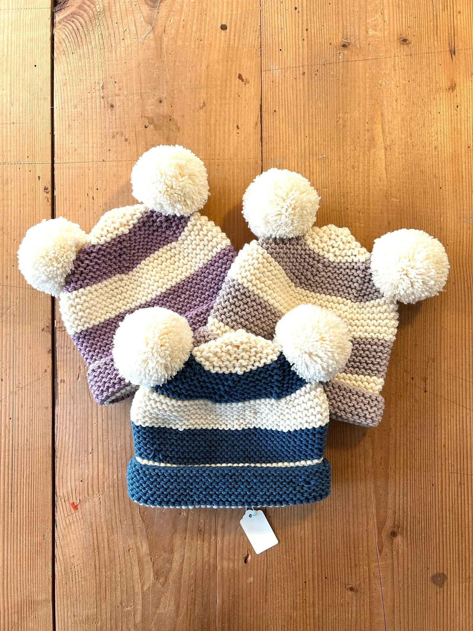 Tre cappelli in maglia a righe larghe, lilla, tortora e blu petrolio con crema, ognuno con due pompon crema