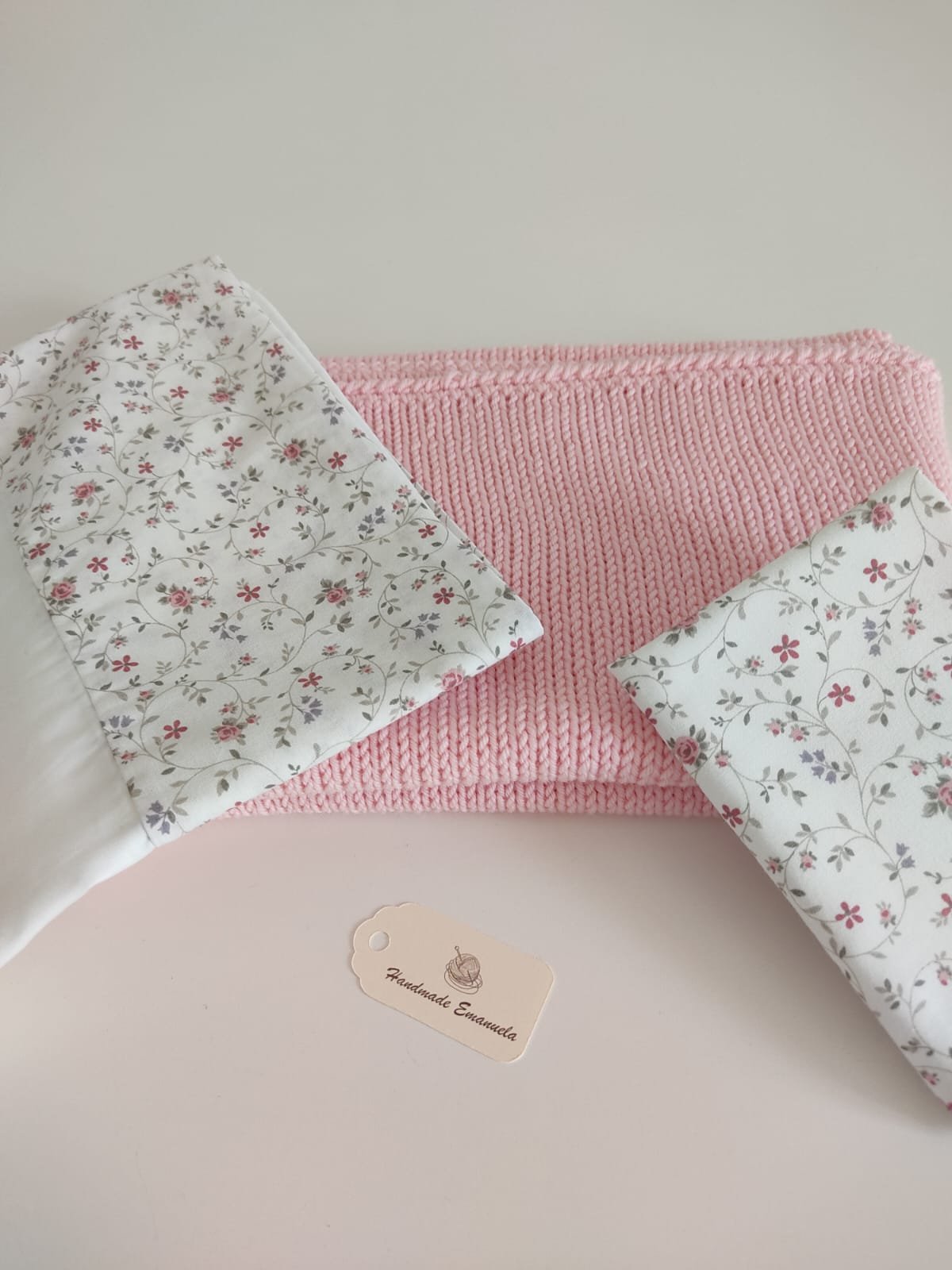 Lenzuolino e federa in cotone bianco a fiorellini rosa piegati su una coperta in maglia rosa, con cartellino