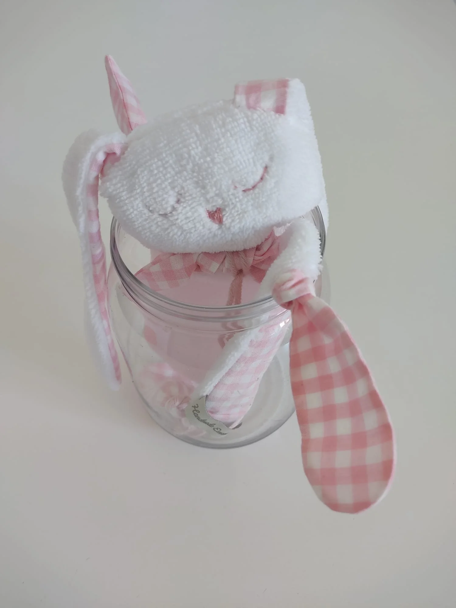 Doudou coniglietto in spugna bianca con orecchie e nodi a quadretti rosa, appoggiato all'imboccatura di un barattolo di vetro