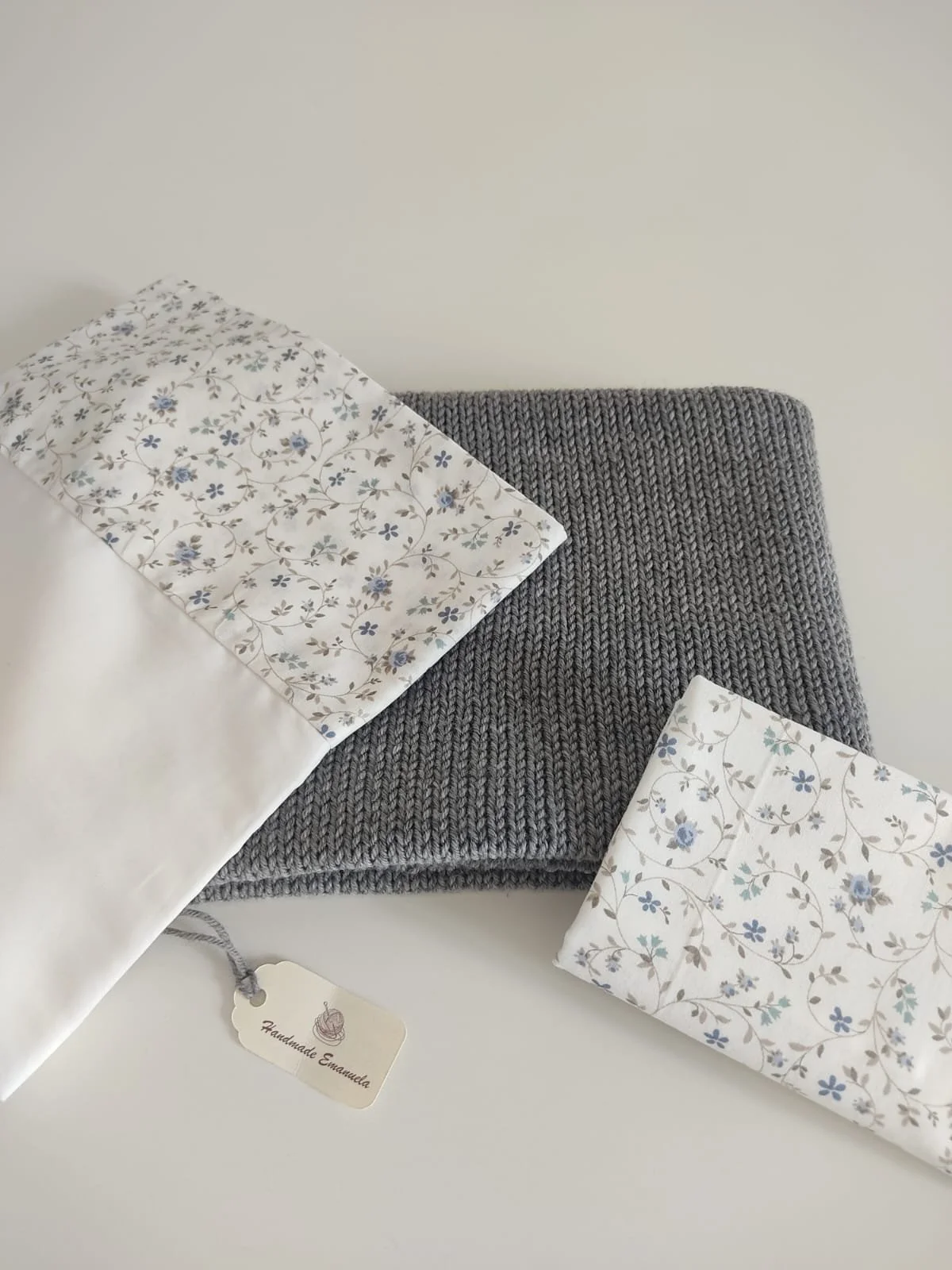 Lenzuolino e federa in cotone bianco a fiorellini azzurri piegati su una coperta in maglia grigia, con cartellino