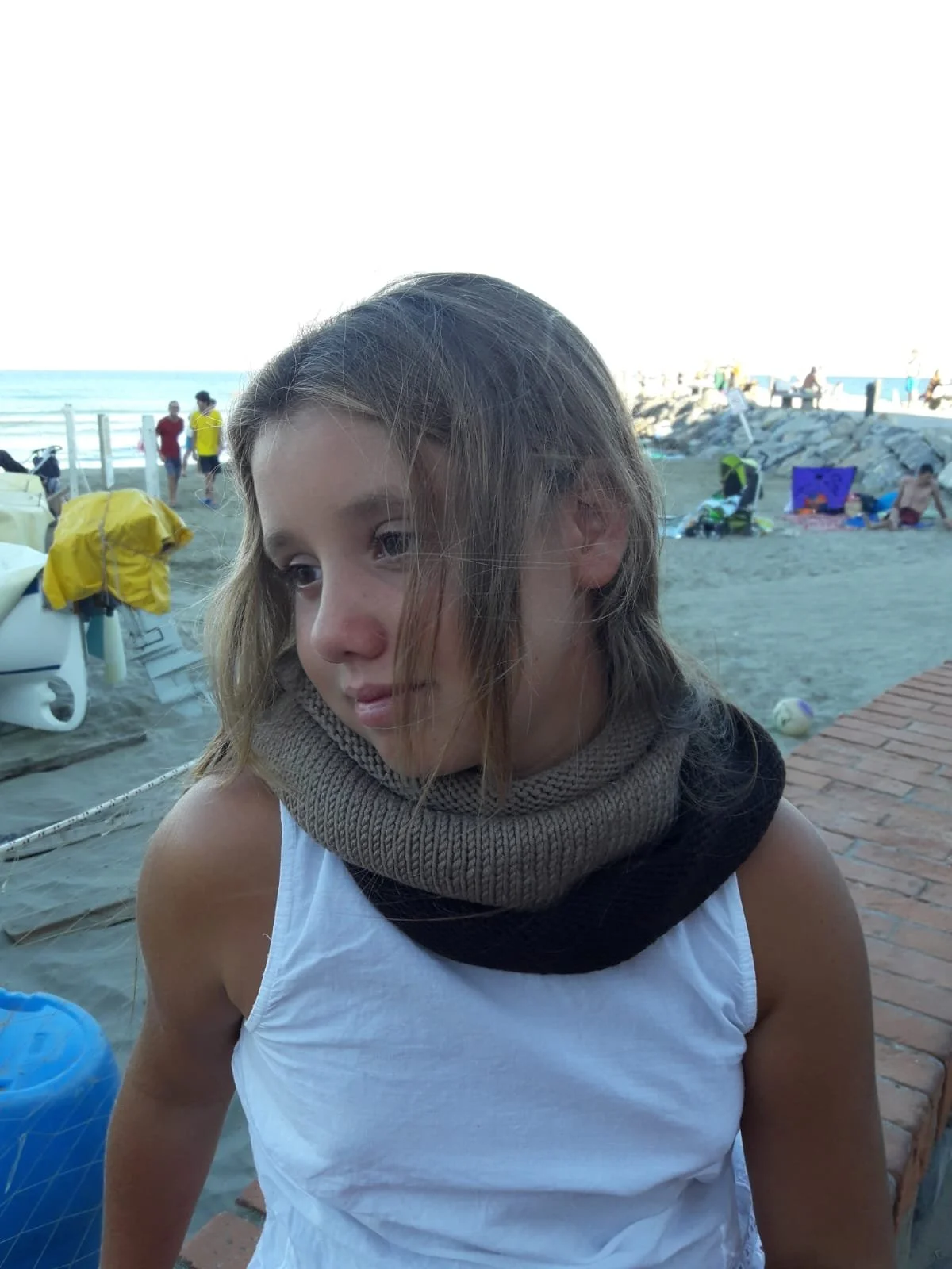 Bambina sulla spiaggia con scaldacollo in maglia beige e marrone scuro avvolto due volte attorno al collo