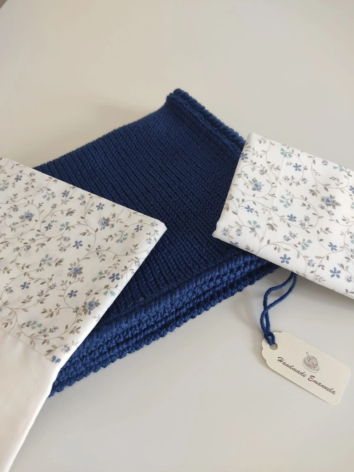 Lenzuolino e federa in cotone bianco a fiorellini azzurri piegati su una coperta in maglia blu, con cartellino