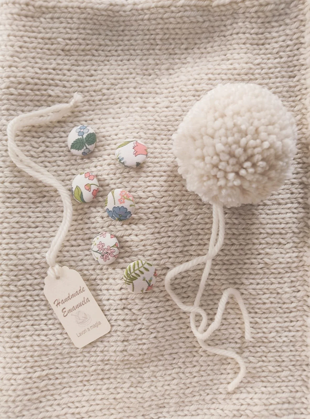 Sei bottoni rivestiti in tessuto fiorito e un grosso pon pon crema su maglia color crema, con cartellino