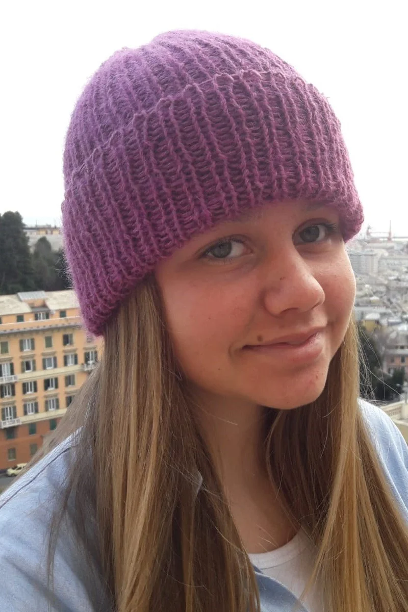 Primo piano di una ragazza con un cappello in maglia lilla a coste con risvolto, con la città sullo sfondo