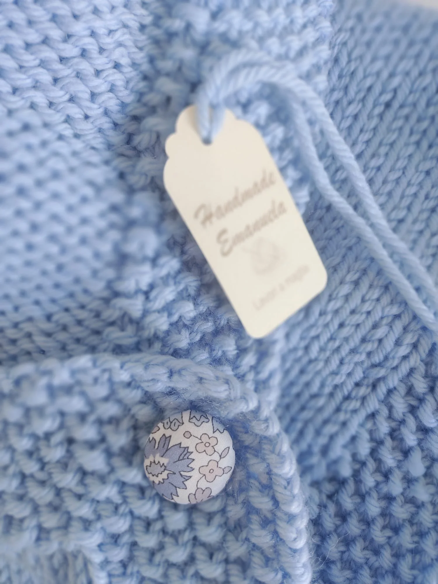 Dettaglio di un bottone rivestito in tessuto fiorito su maglia azzurra, con cartellino di cartoncino e filo