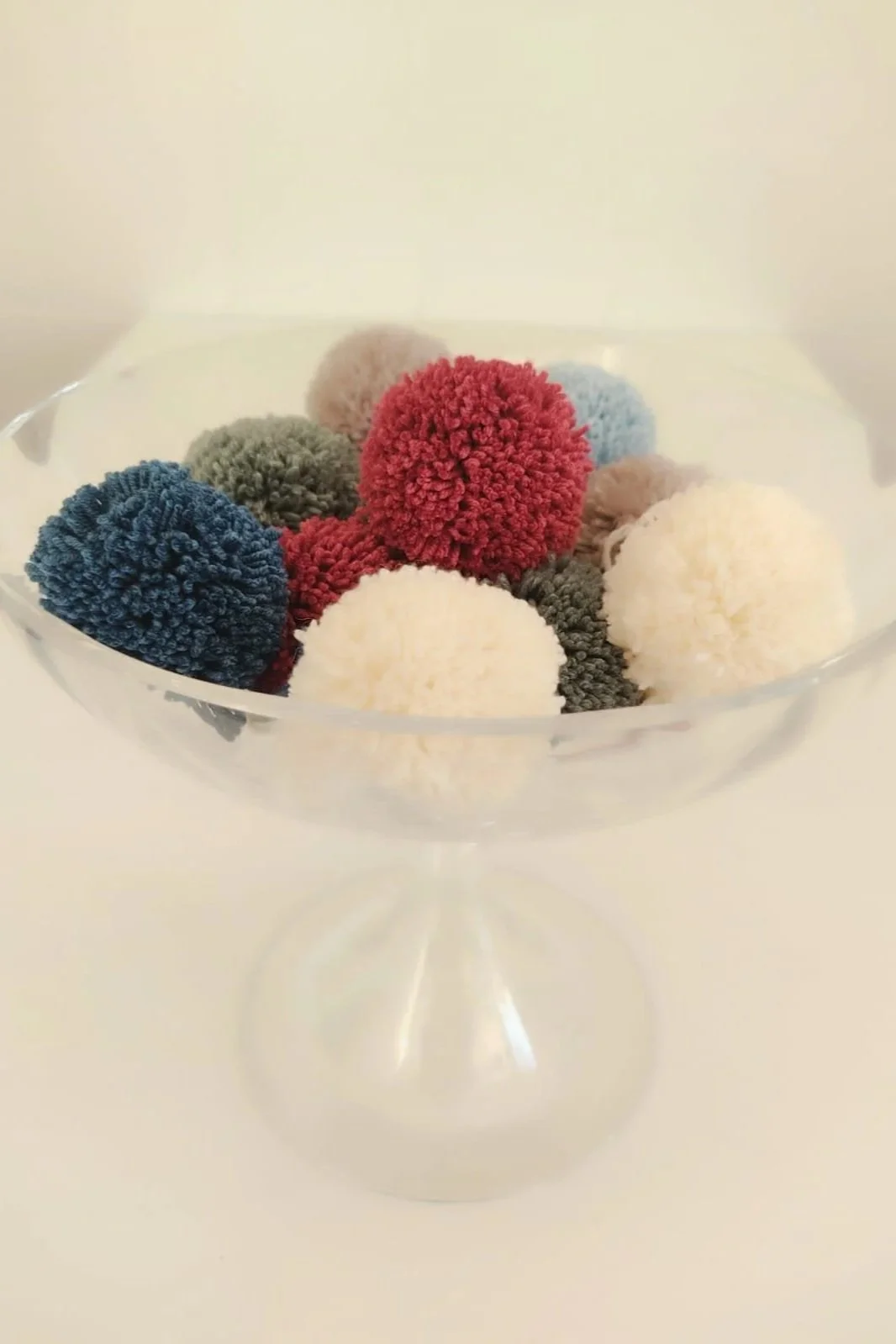 Pon pon in lana di colori diversi, blu, bordeaux, panna, grigio e azzurro, in una coppa di vetro trasparente