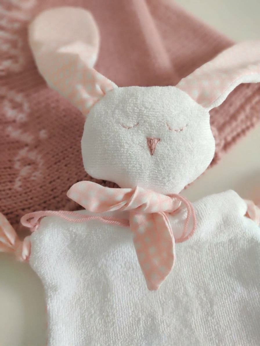 Primo piano di un doudou coniglietto in spugna bianca con orecchie rosa a fantasia, su una coperta in maglia rosa antico